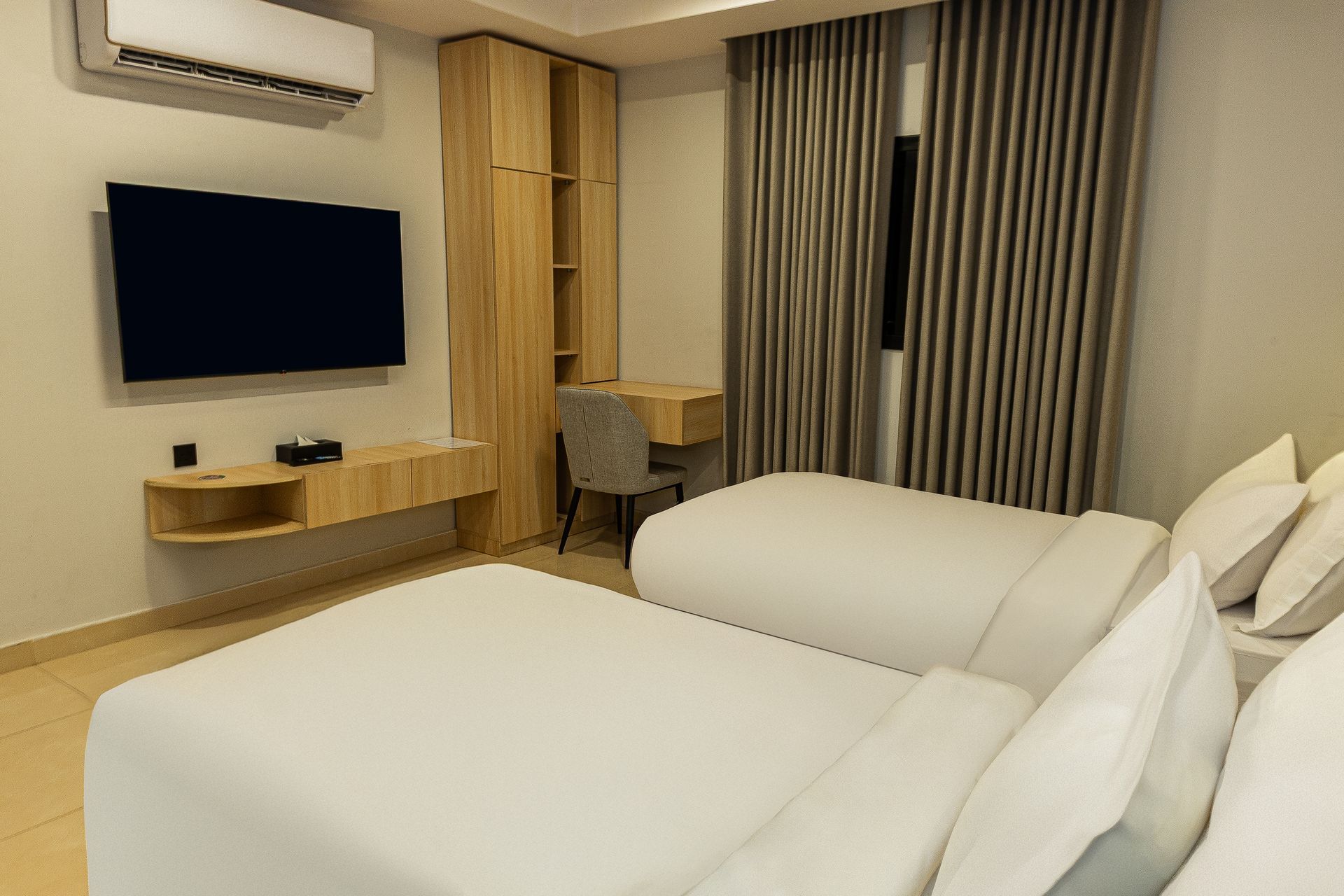 Modern hotel room with twin beds, wall-mounted TV, desk, air conditioner, and floor-to-ceiling curtains