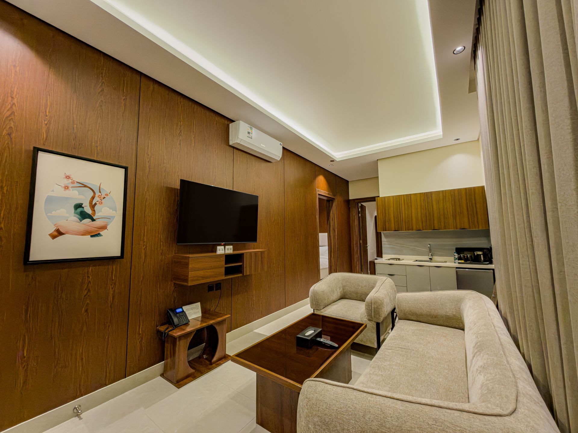 Modern studio apartment with wood-paneled walls, a mounted TV, a beige sofa and armchair, and a small kitchenette.