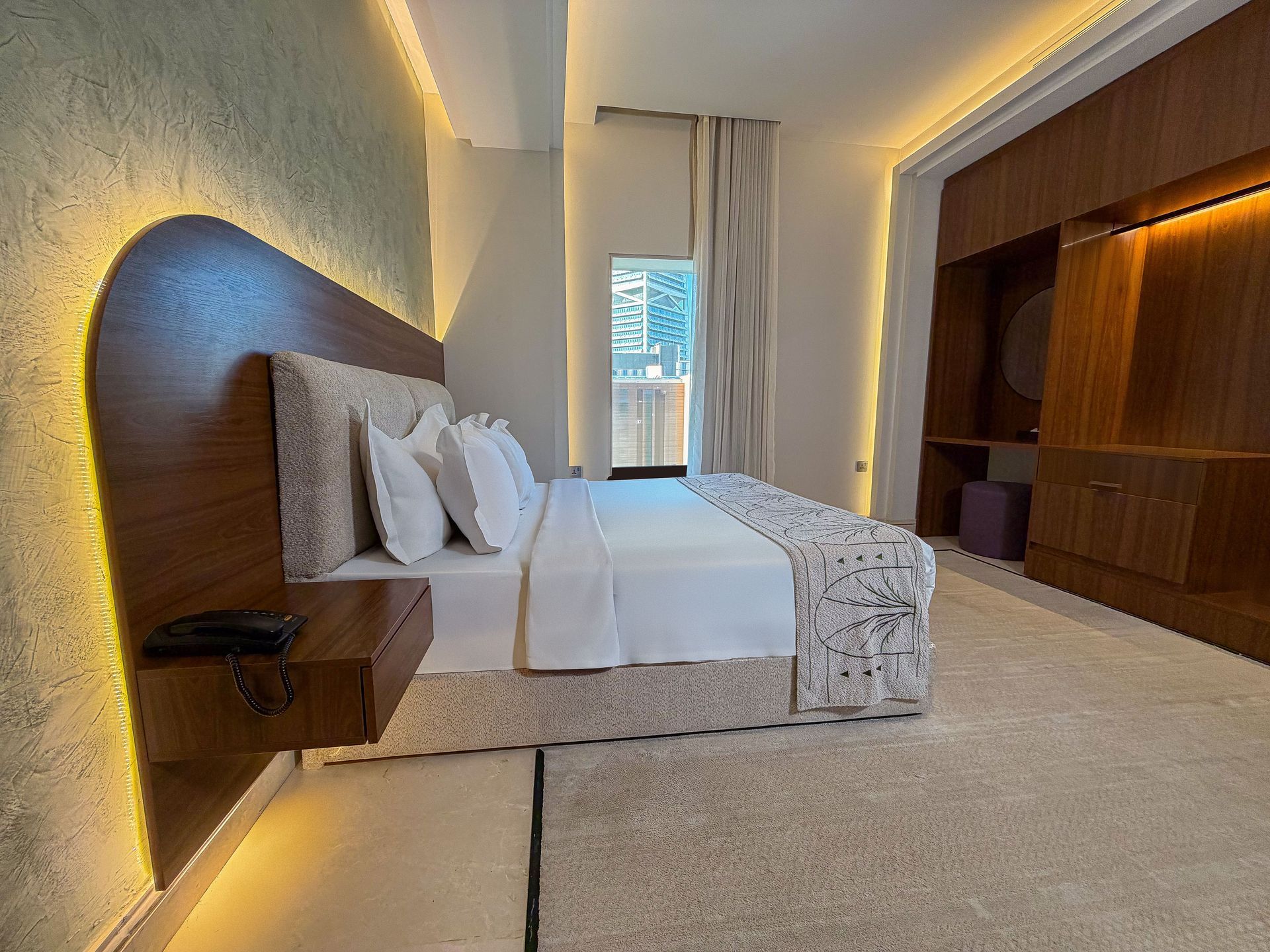 Modern bedroom with a neatly made bed, warm wood accents, and a window with light curtains.