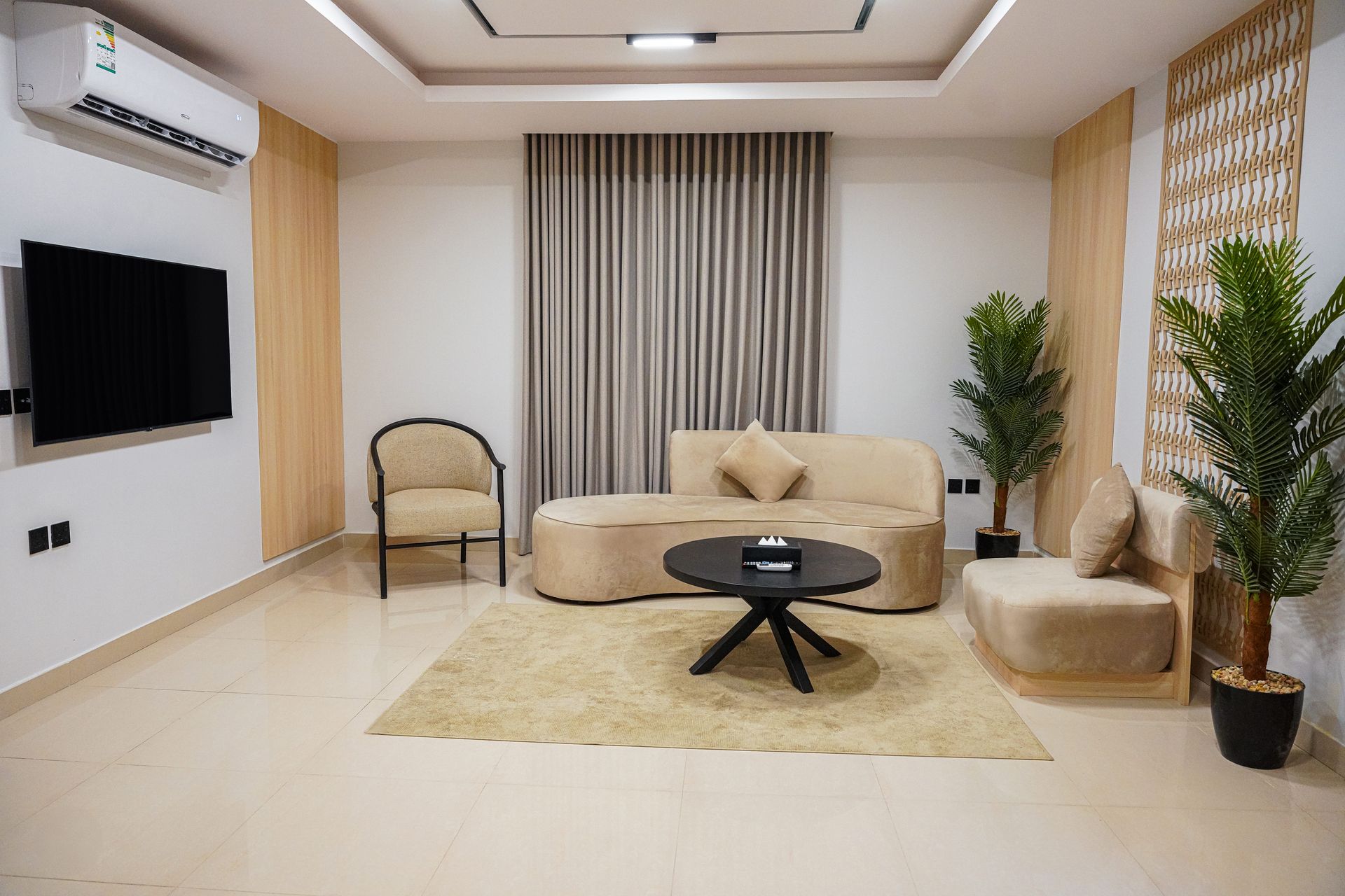 Modern living room with beige sofa, wall-mounted TV, curtains, plants, and a coffee table on a rug.