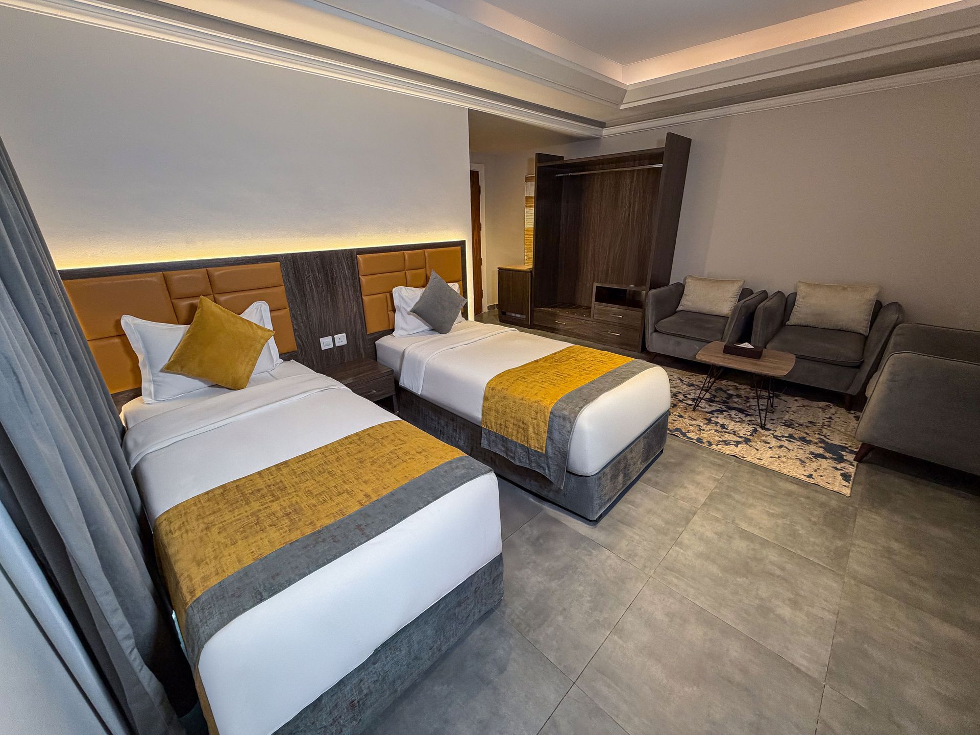 Modern hotel room with two twin beds, gray sofa, wood accents, and warm ceiling lighting