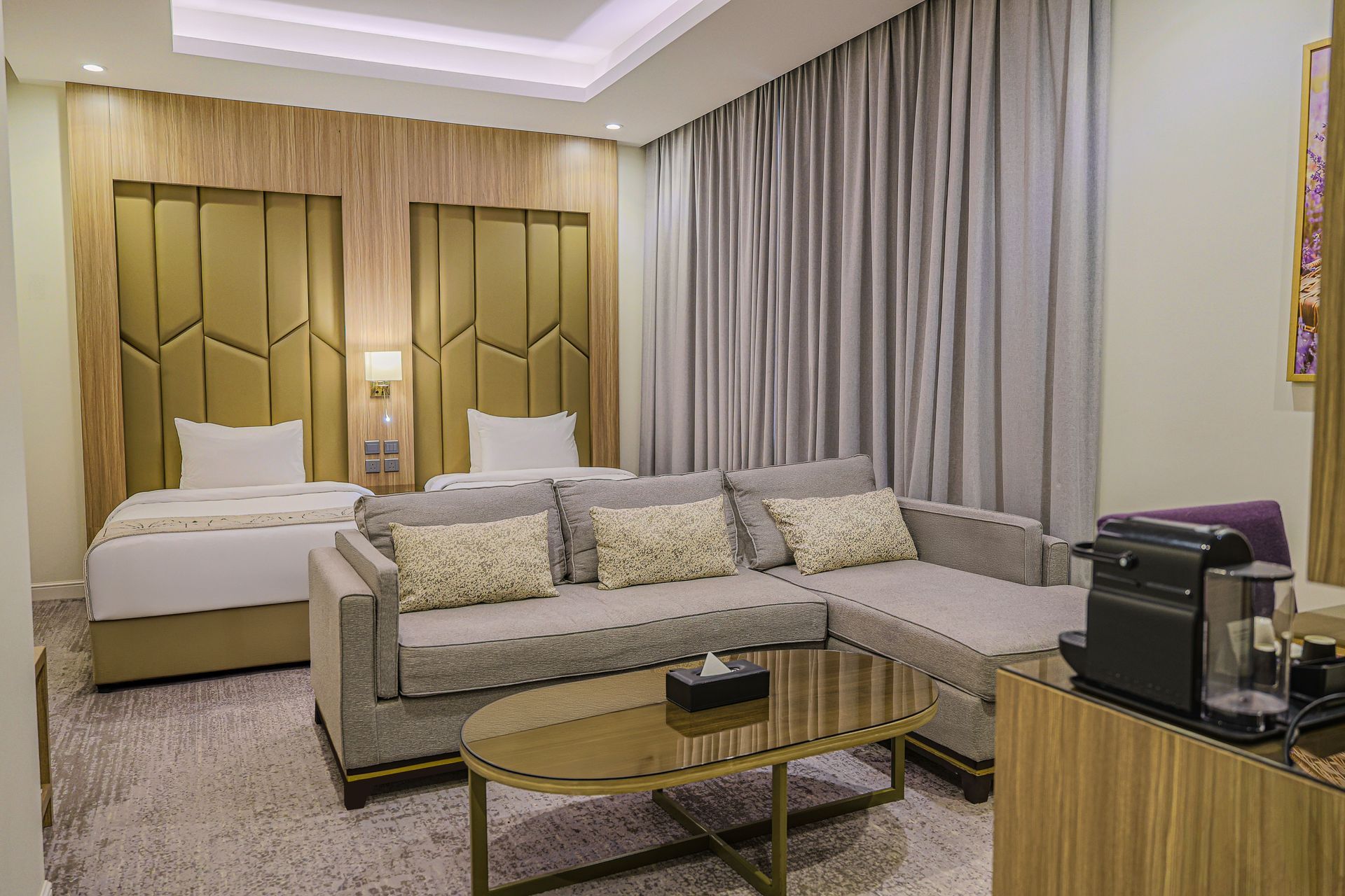 A modern hotel room with two twin beds, a grey sectional sofa, an oval coffee table, and soft, neutral lighting.