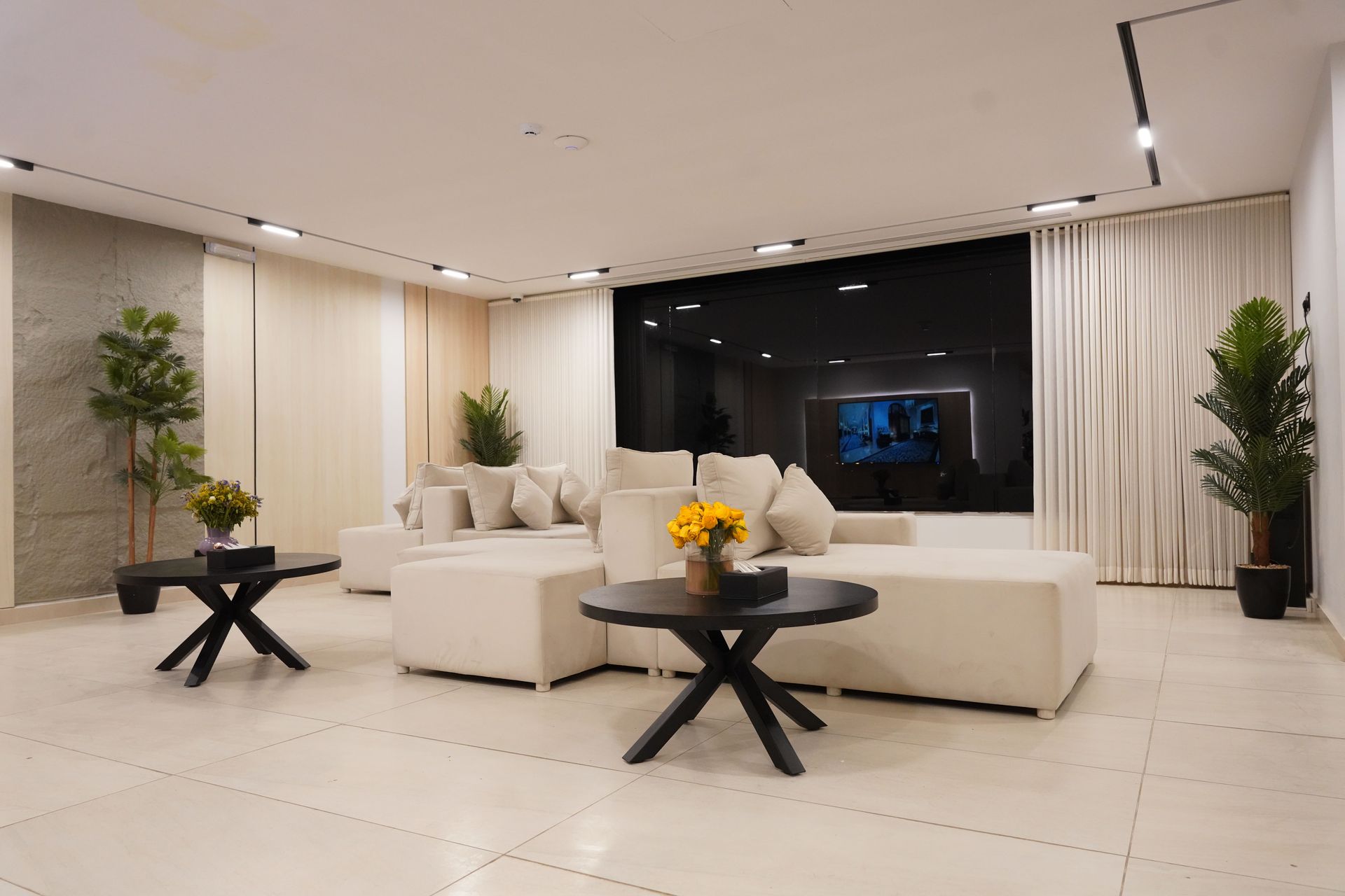 Modern living room with beige sectional sofas, two black round coffee tables with yellow flowers, and potted plants.