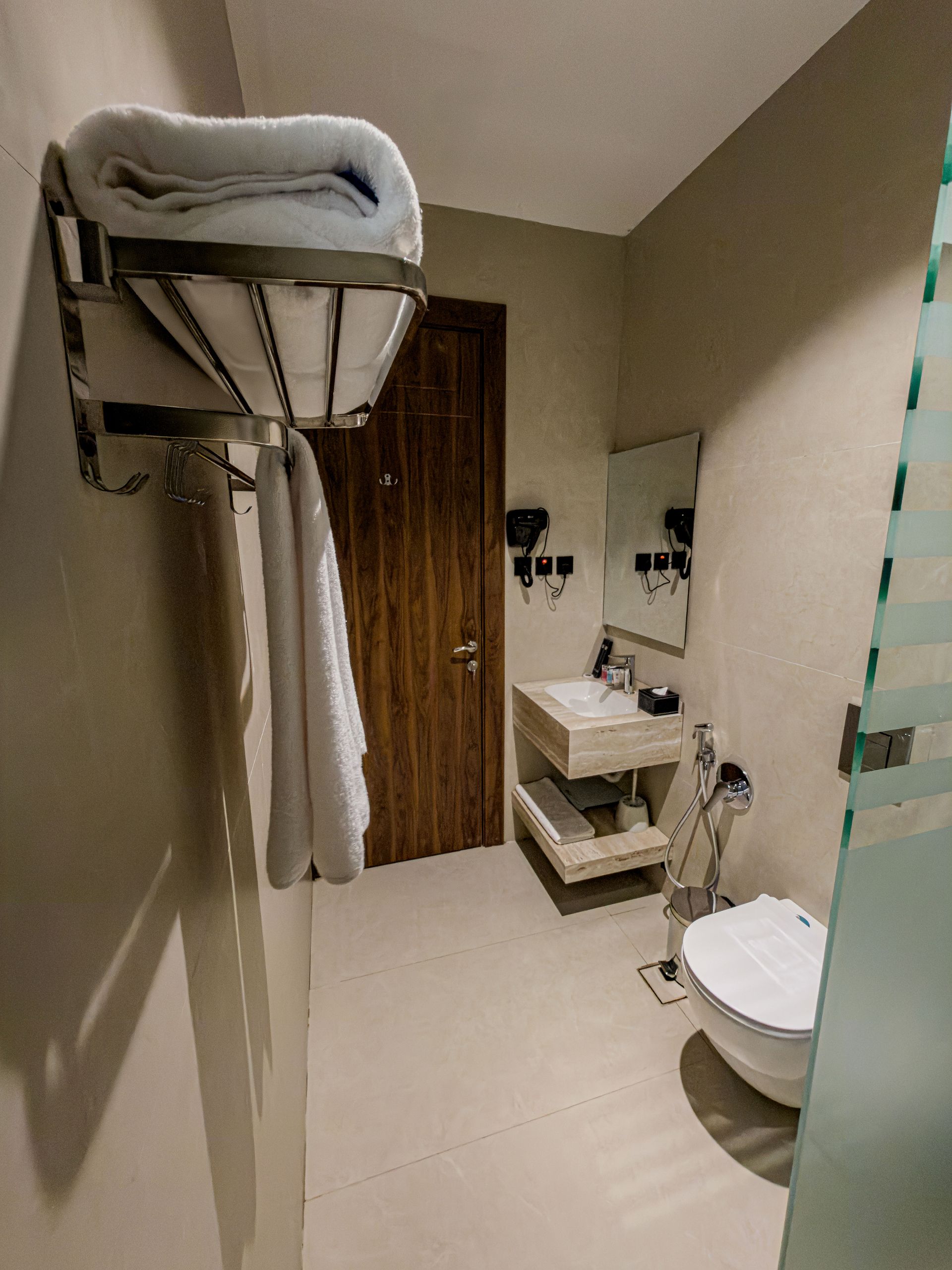 A modern hotel bathroom with a wall-mounted towel rack, a wooden door, a floating vanity with a sink, and a white toilet.