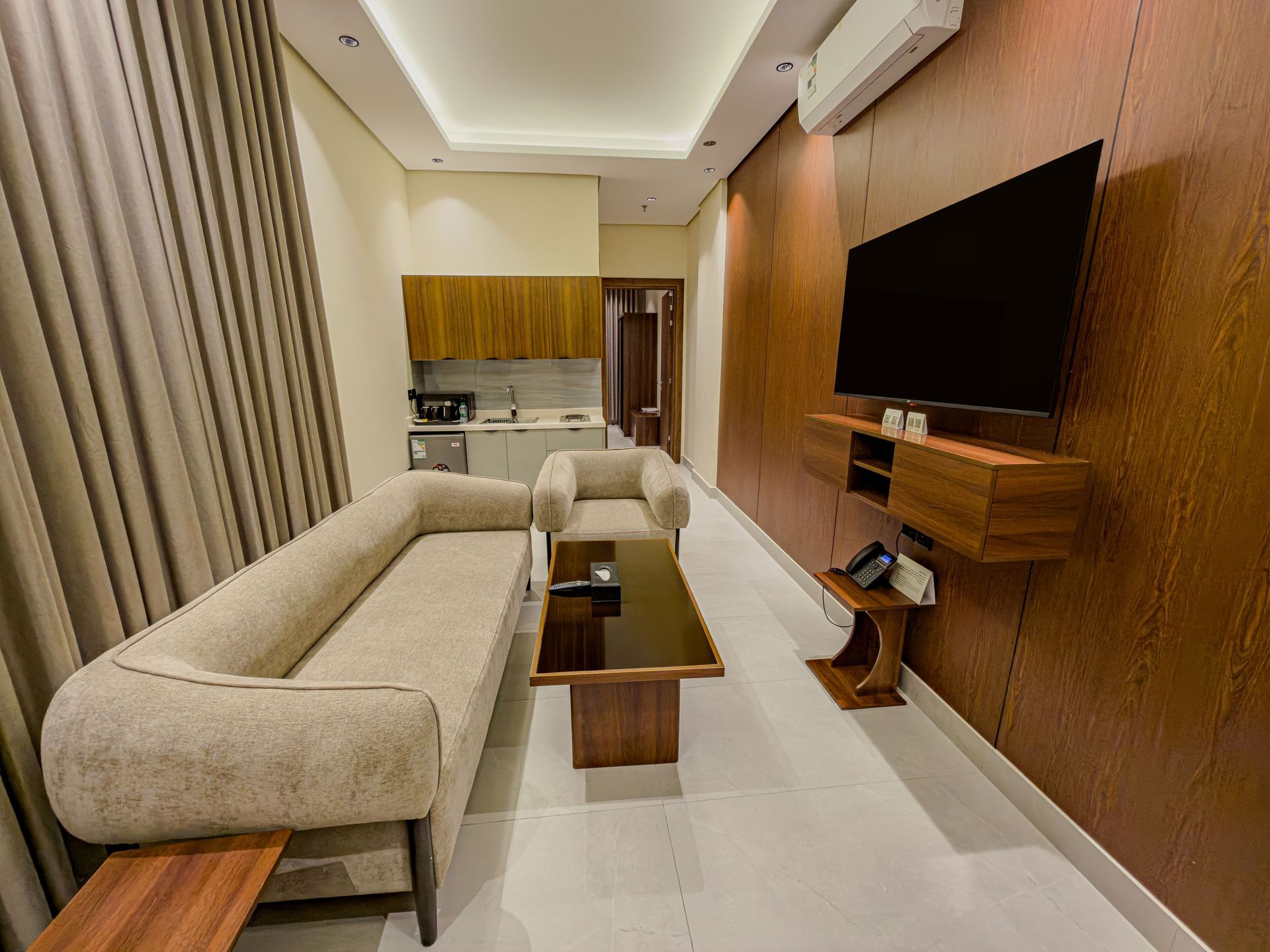 A modern living room with a beige sofa, a coffee table, a single armchair, and a wall-mounted TV on wood-paneled wall.