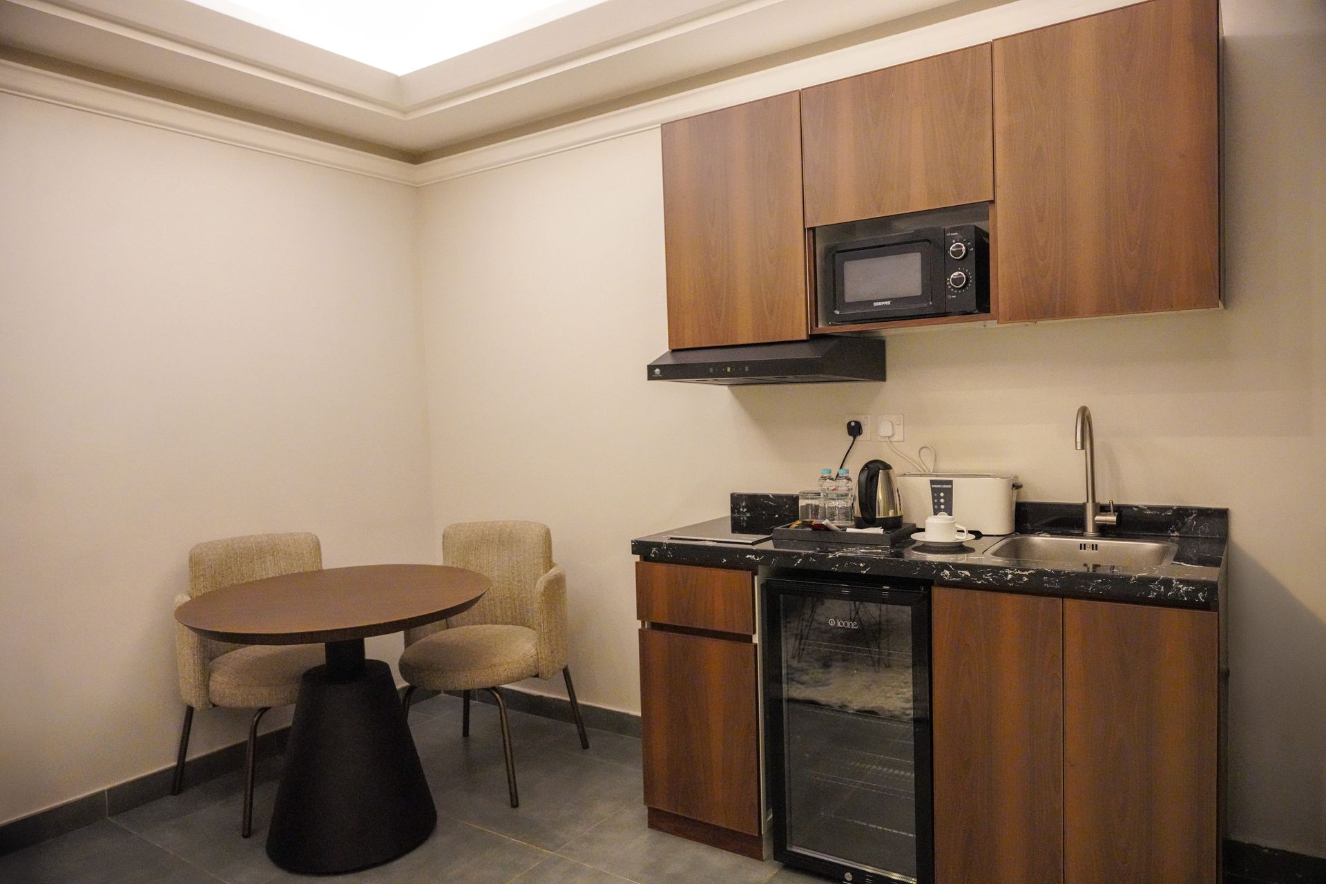 Small hotel room kitchenette with wood cabinets, mini fridge, microwave, sink, and a round table with two chairs.