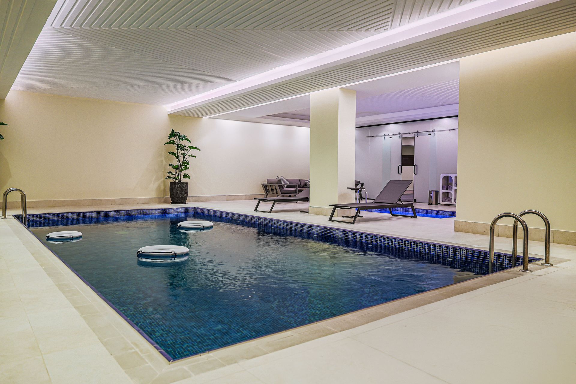 An indoor swimming pool with blue water and metal ladders, beside a modern lounge area with dark sofas and chairs.