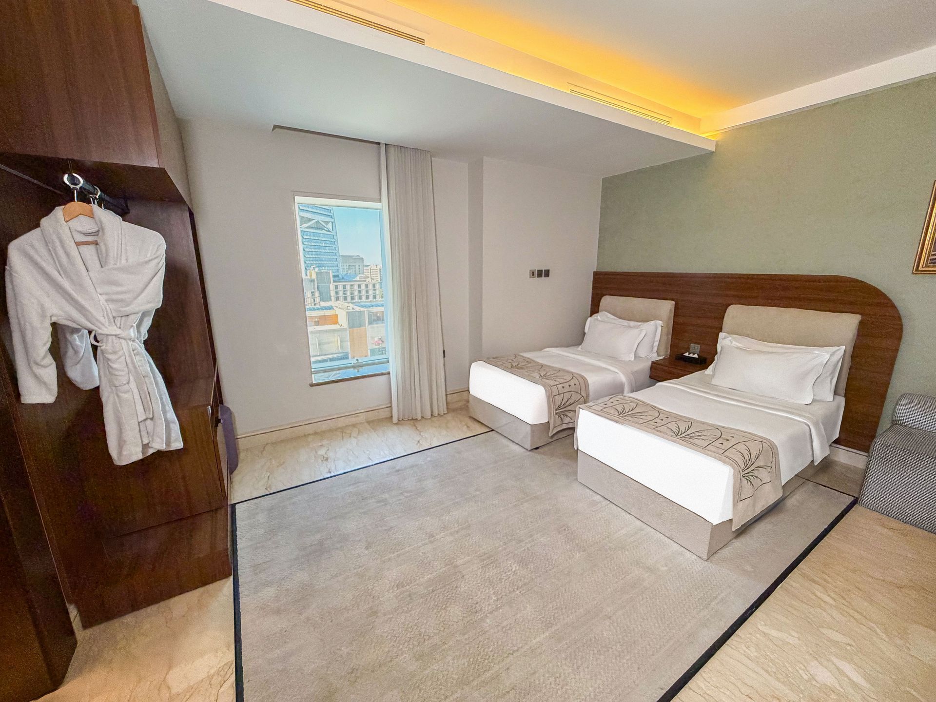 Modern hotel room with two single beds, warm lighting, and a small window view of city buildings
