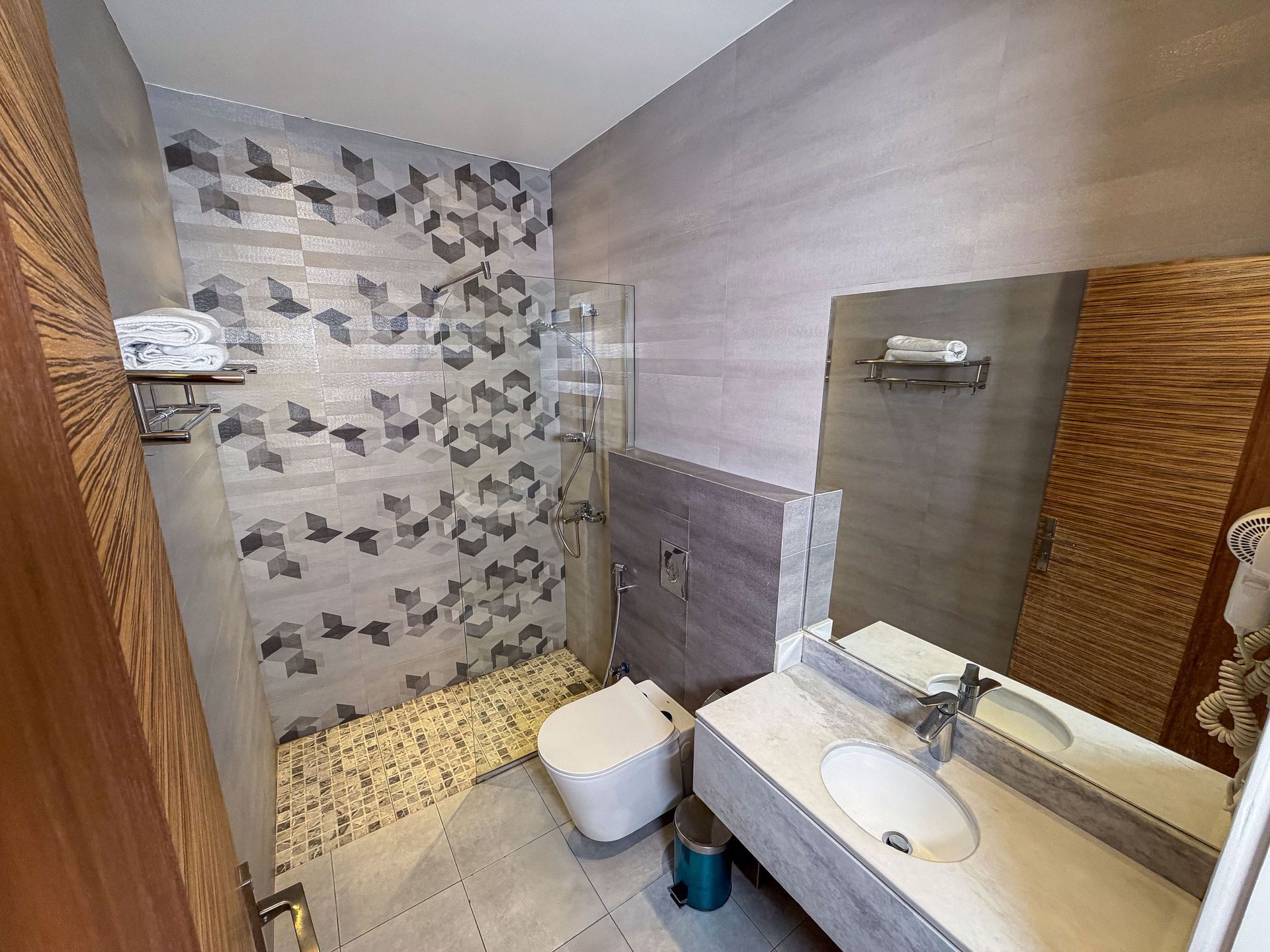 Small bathroom with stone-pattern shower wall, wall-mounted toilet, sink, mirror, and wicker hamper