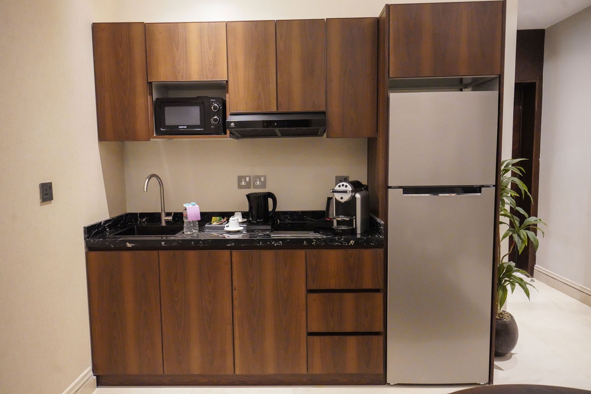 Compact kitchenette with wood cabinets, black countertop, sink, microwave, coffee maker, and silver refrigerator.