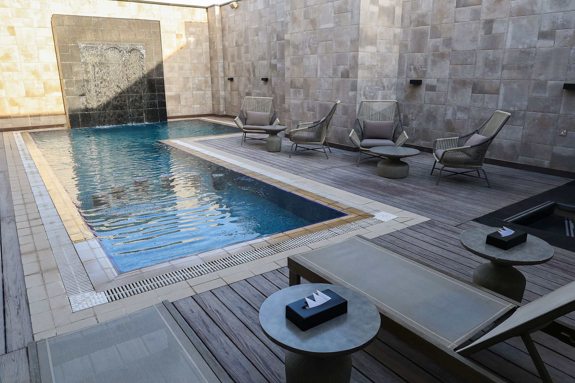 An outdoor rectangular swimming pool with a stone wall water feature, loungers, and side tables on a wooden deck.