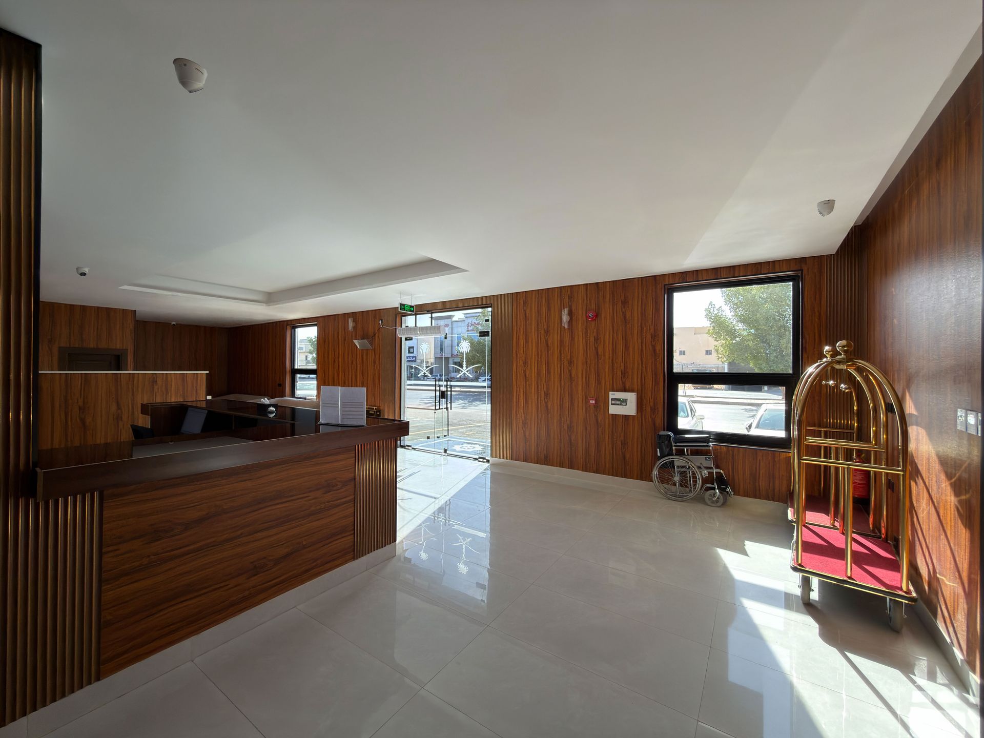 A hotel lobby with wood-paneled walls, a reception desk, a luggage cart, a wheelchair, and a glass front entrance.