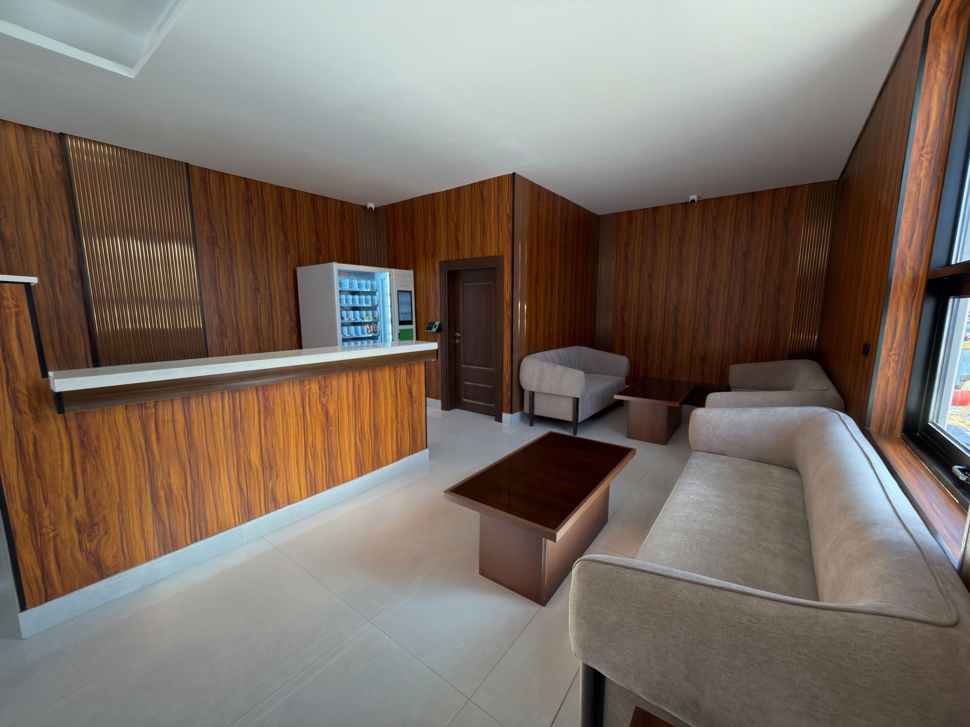A hotel lobby featuring wood-paneled walls, a reception counter with a vending machine, and beige couches around a table.