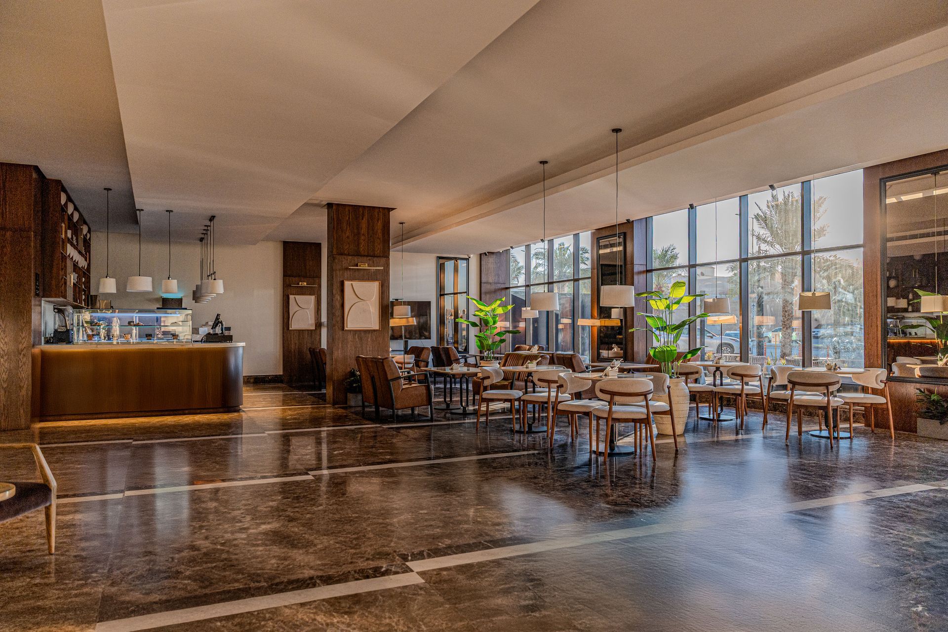 An elegant hotel lobby featuring marble floors, wooden pillars, a coffee bar, and multiple tables with chairs by windows.