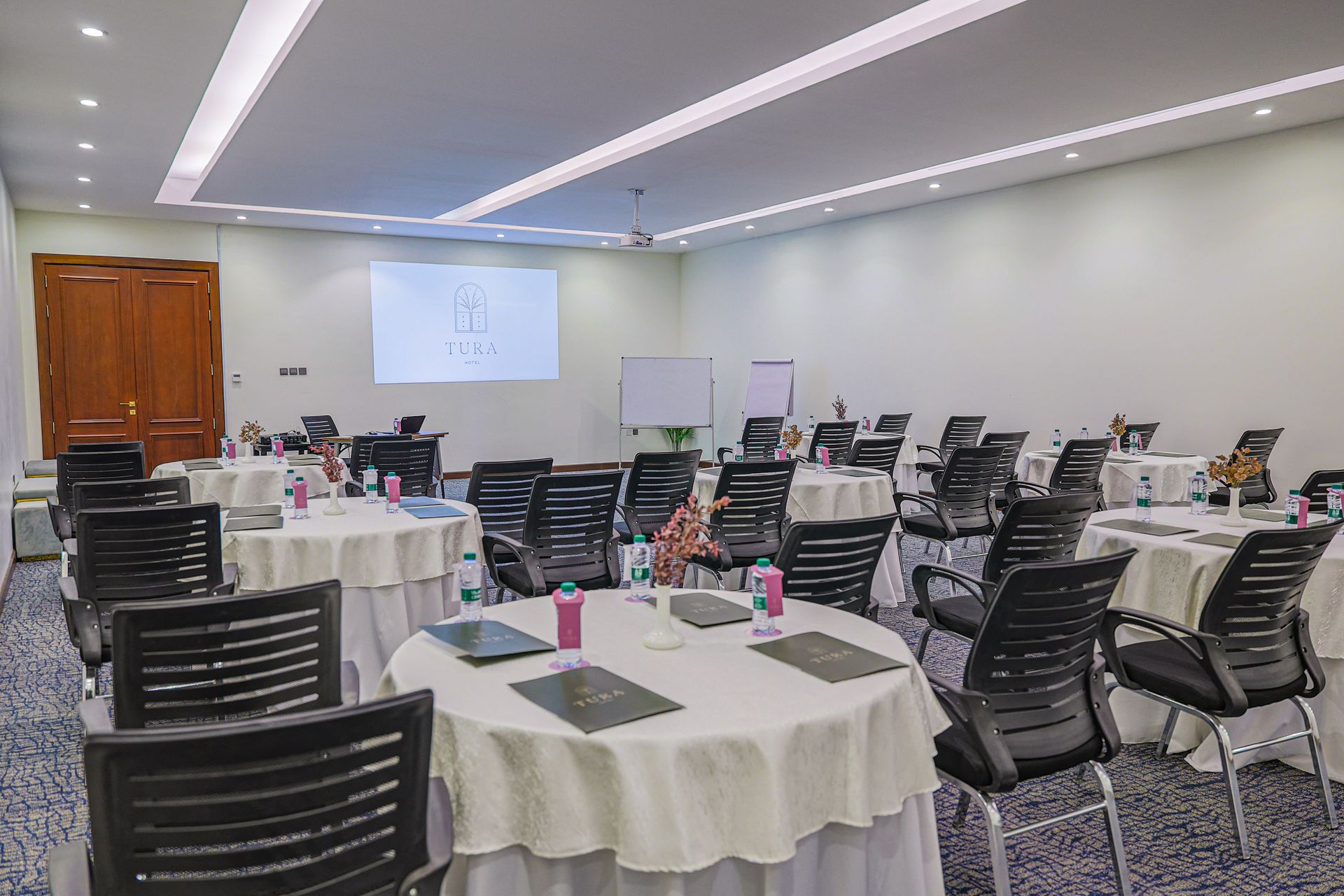 A conference room set with several round tables covered in white cloths, surrounded by black chairs and a projector screen.