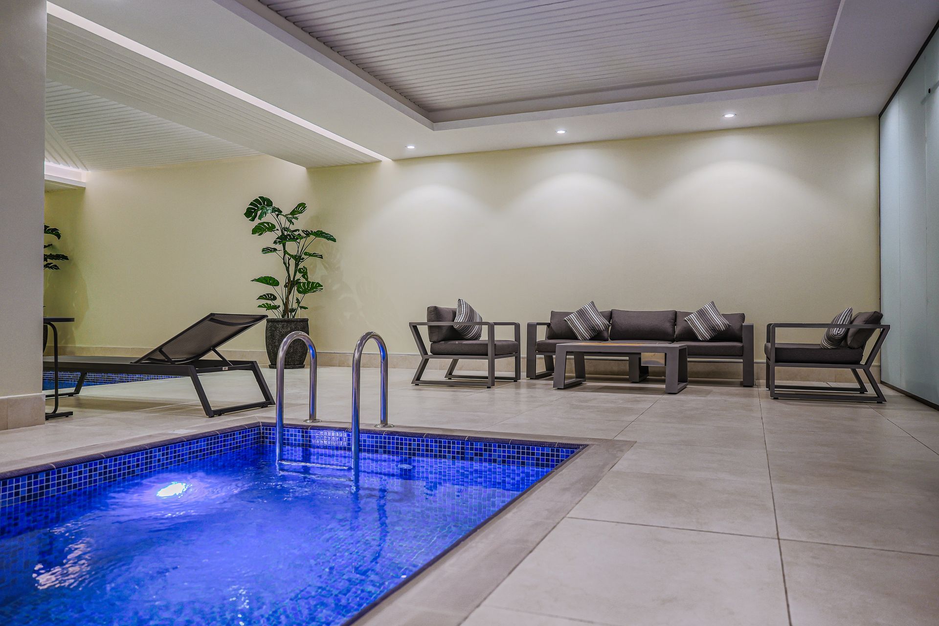 An indoor pool area featuring a blue-lit rectangular pool, a lounge chair, and modern outdoor seating set on tile floors.