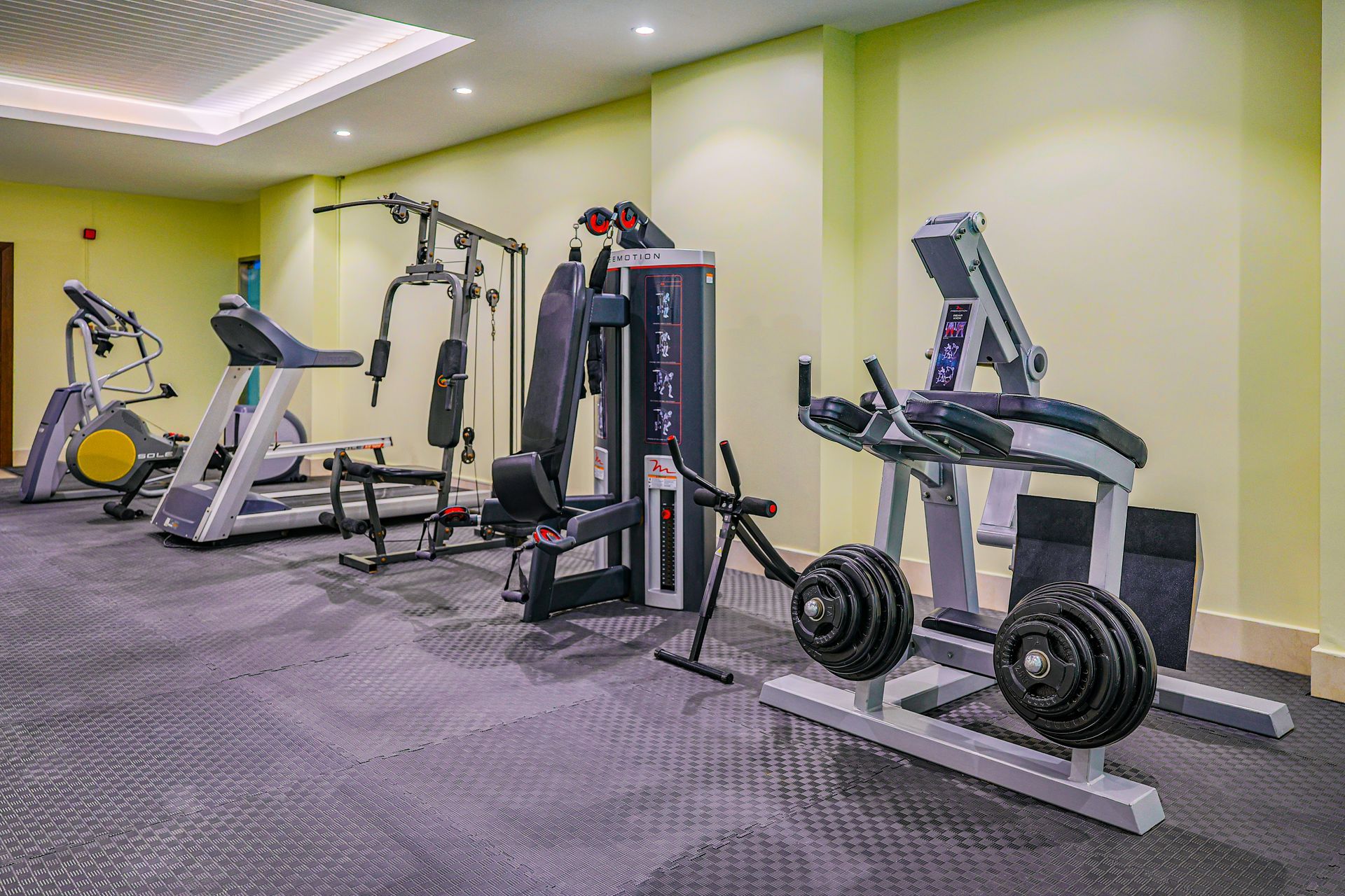 An indoor gym with various workout equipment, including a treadmill, a cable machine, and a bench with weight plates.