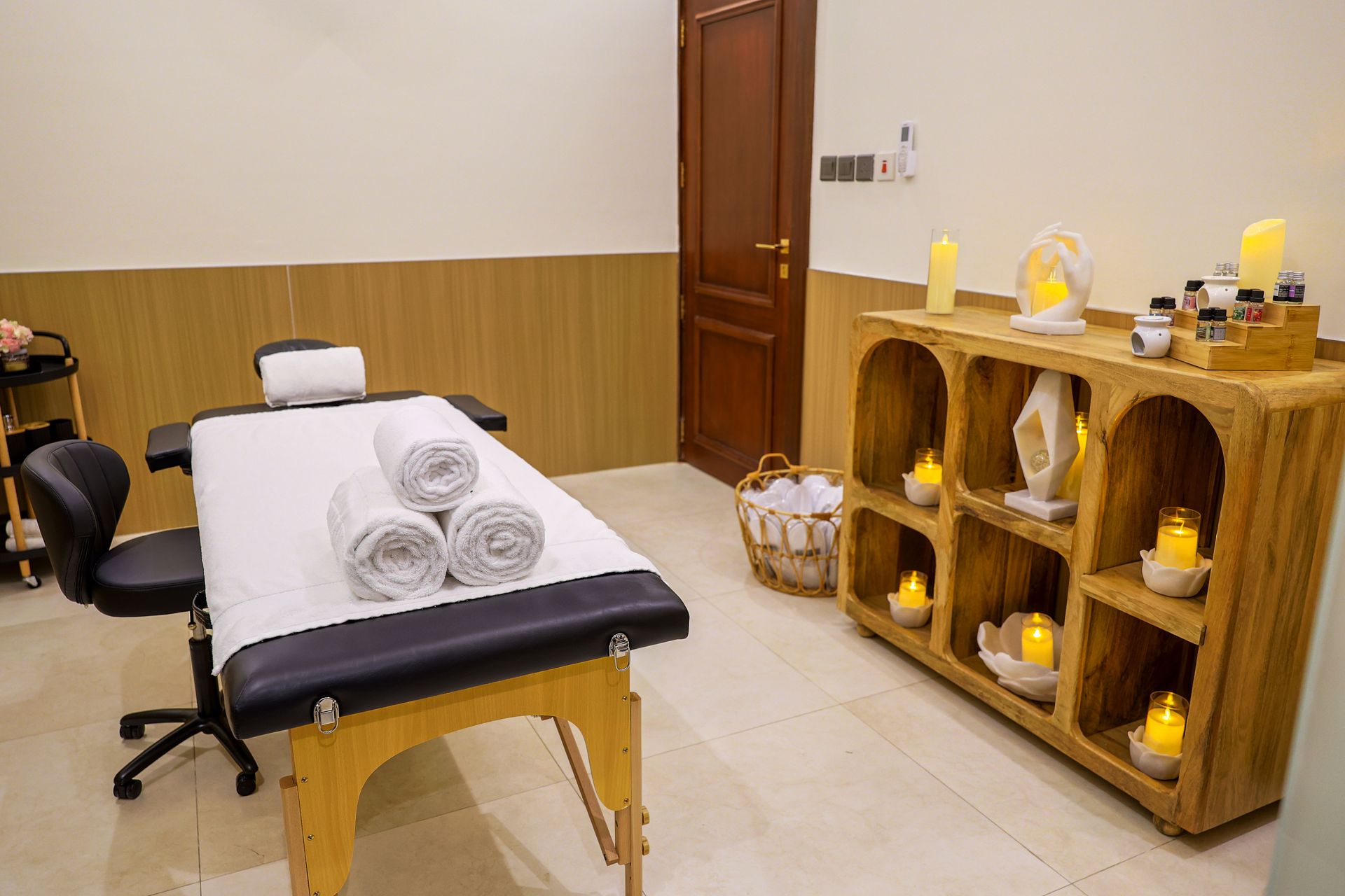 A massage treatment room with a therapy bed, a chair, and a wooden shelf displaying candles and spa supplies.
