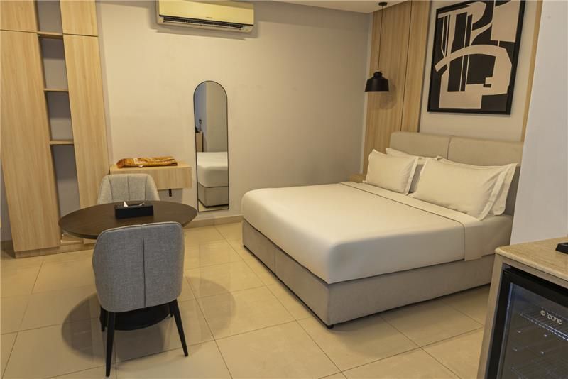 Modern hotel room with a white bed, chair, small table, mirror, and abstract wall art.