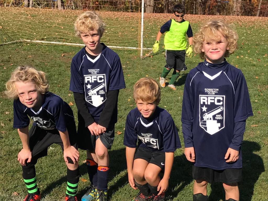 Four children in navy soccer uniforms posing on a grassy field, with a goal and autumn trees behind them