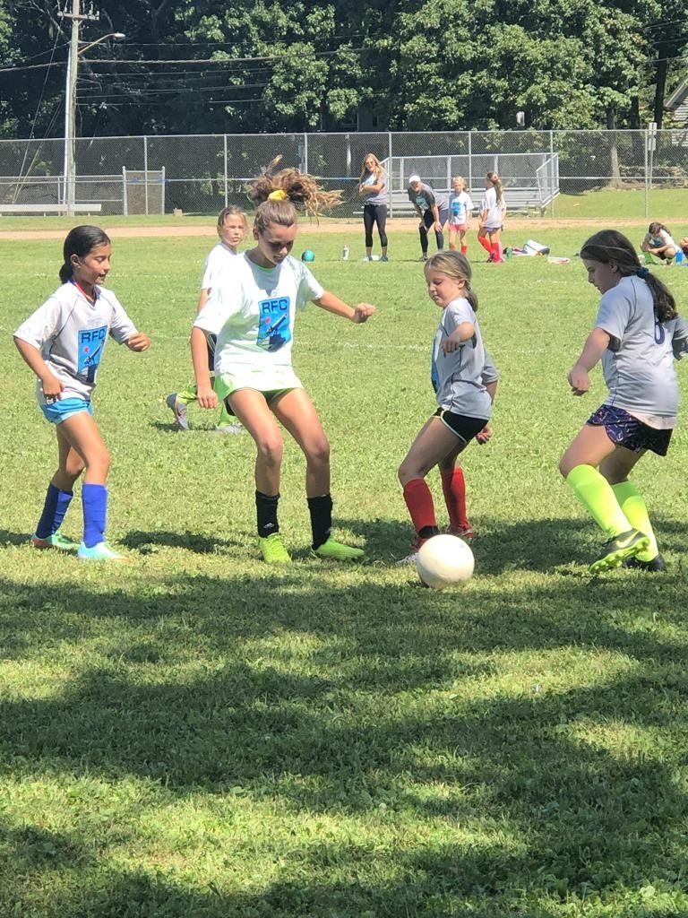 Youth soccer players chase a ball on a sunny grassy field near a fence.