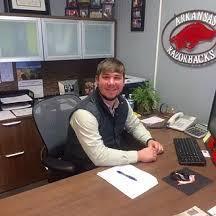 Man sitting at an office desk, smiling beside Arkansas Razorbacks wall sign.
