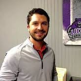 Smiling man in a gray pullover standing indoors beside a purple and white sports sign