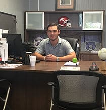 Man seated behind a desk in an office, smiling with a football helmet and framed items on shelves behind him