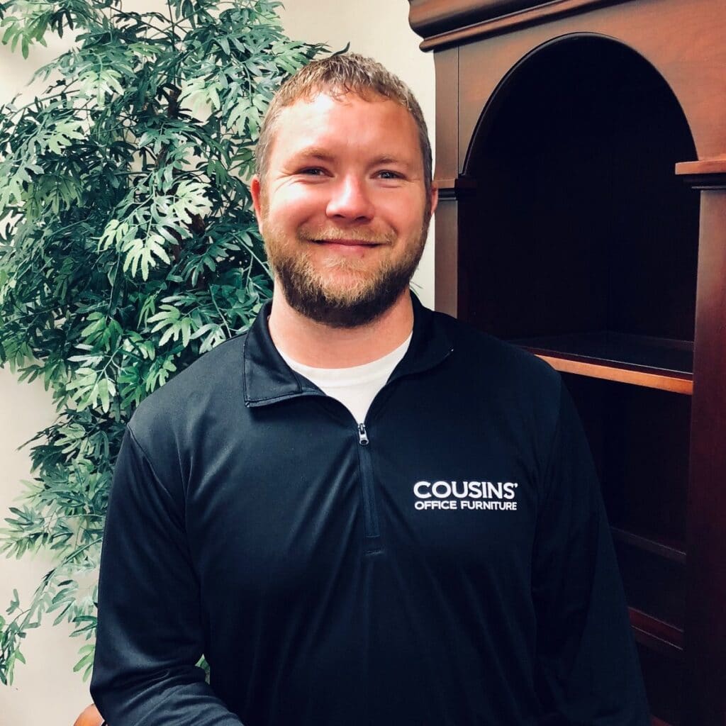 Smiling person in a black Coussins shirt standing beside an indoor wooden archway and leafy plant