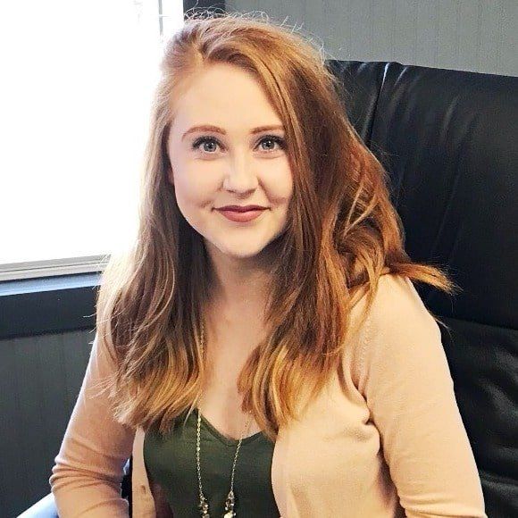 Smiling woman with long hair seated in a black chair, wearing a beige cardigan and green top