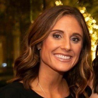 Smiling woman with wavy brown hair against a warm, blurred background with lights