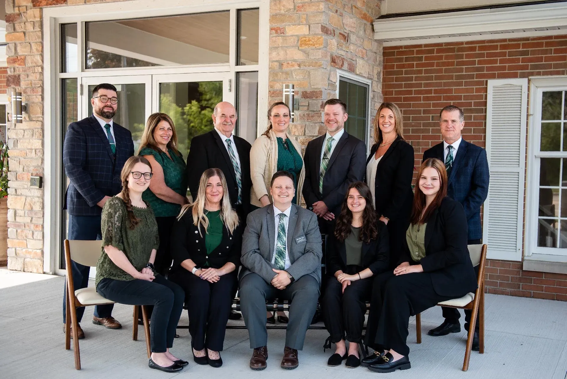 Group portrait of eleven professionals in business attire standing and sitting outside a brick building.