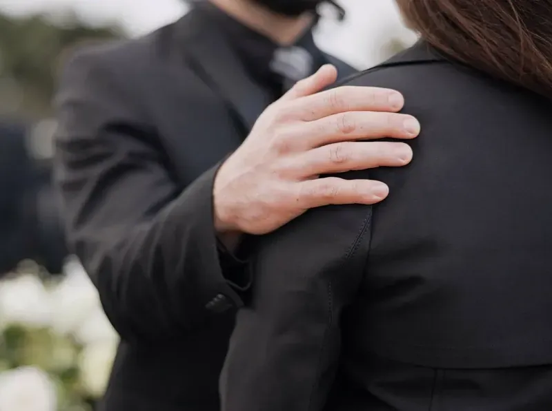 A person in a black suit places a hand gently on the shoulder of another person wearing a black blazer.
