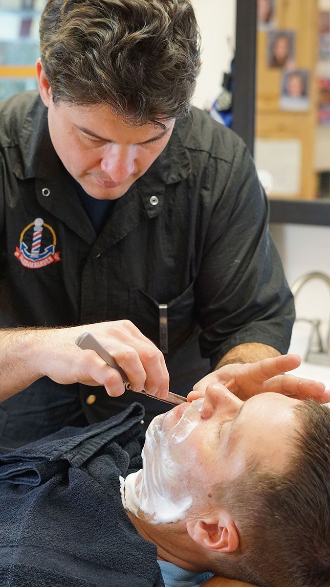 A man lying back in a barber chair while a professional barber gives him a traditional wet shave with a straight razor.