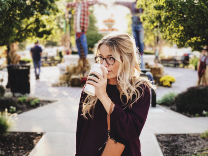 A woman with glasses in a dark sweater drinks from a coffee cup outdoors in a park with scarecrows in the background.