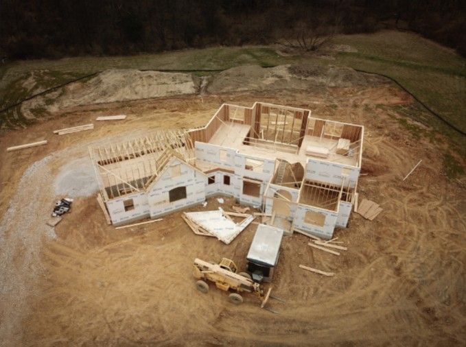 An aerial view of a residential house under construction, with exposed wooden framing and foundation on a dirt lot.