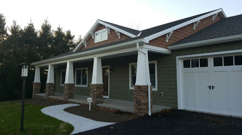 A single-story house with green siding, a brick gable, a stone-pillared porch, and an attached white garage.