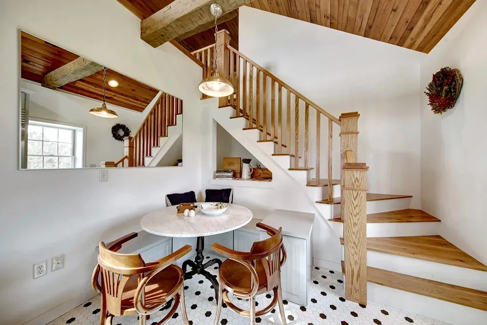 A breakfast nook under a wooden staircase with a marble-topped table, two wooden chairs, and white-and-black floor tiles.