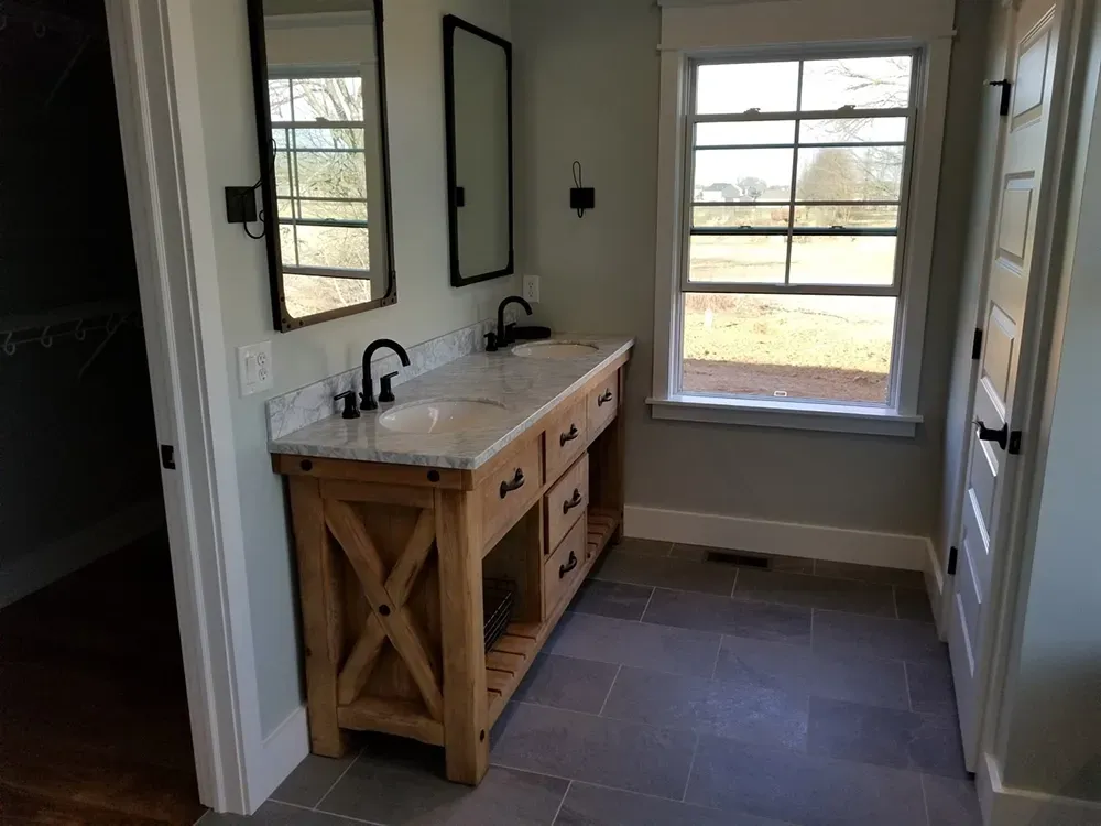 A bathroom vanity with a marble top, rustic wood cabinet, and dual mirrors under a window with grey tiled floors.