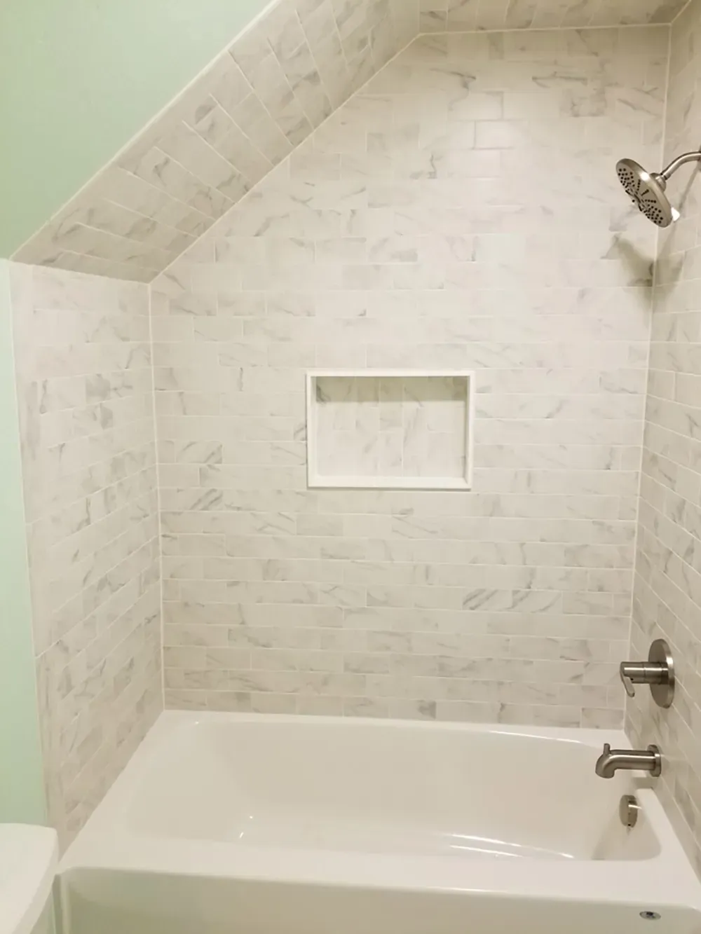 A white bathtub and shower surround with light marble-patterned tiles, a recessed wall niche, and brushed nickel fixtures.