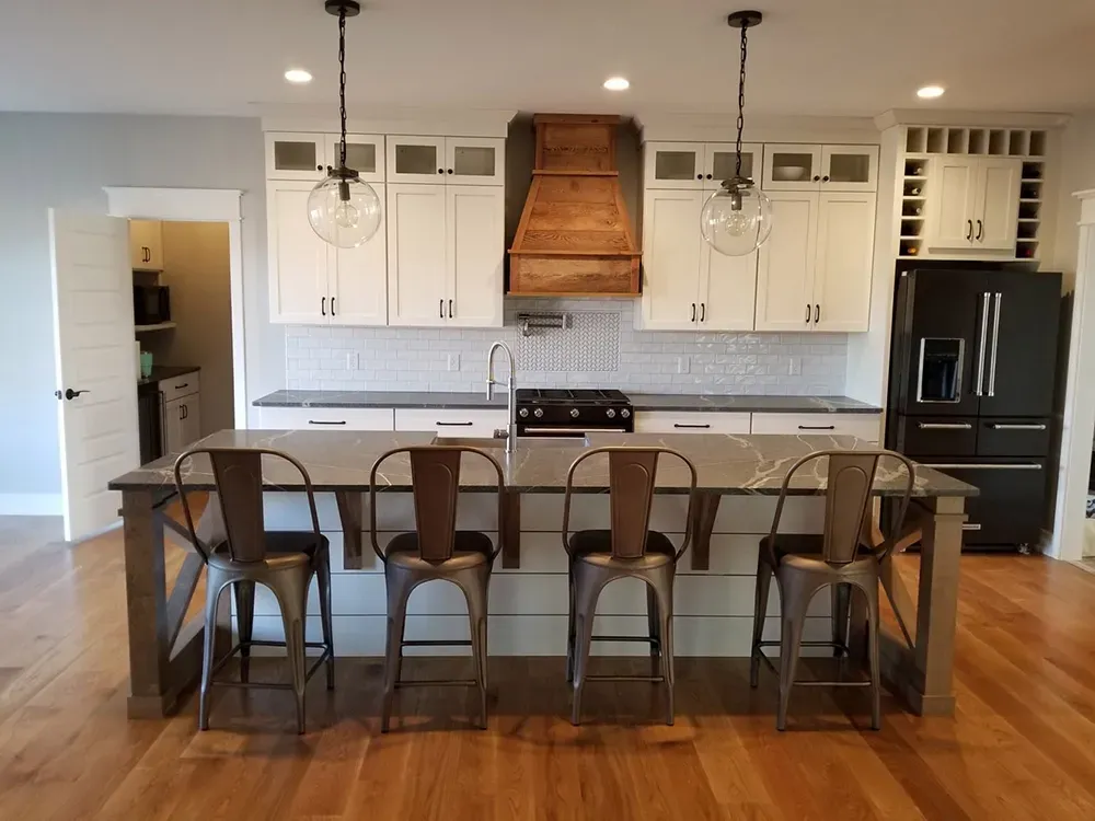 A bright, modern kitchen with white cabinets, a wooden range hood, a large island with four metal stools, and wood floors.