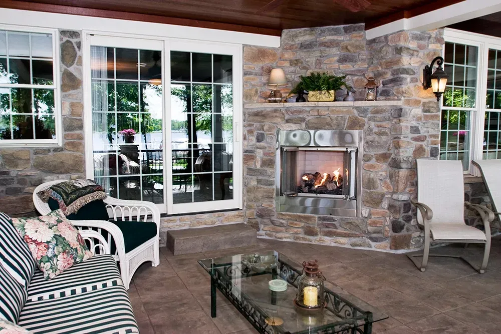 A cozy stone sunroom with a lit fireplace, glass-topped coffee table, and patio furniture facing sliding glass doors.