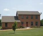 A two-story brick house with a gabled roof and a smaller attached single-story section, set in a grassy yard.