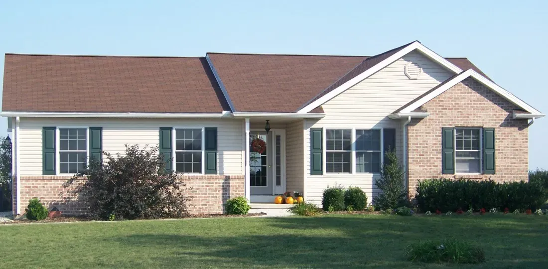 A one-story suburban home with tan siding, a brown shingled roof, brick accents, and green shutters under a clear blue sky.