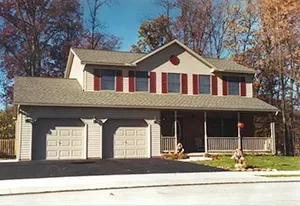 A beige, two-story house with a two-car garage, front porch, and red shutters, surrounded by autumn trees.