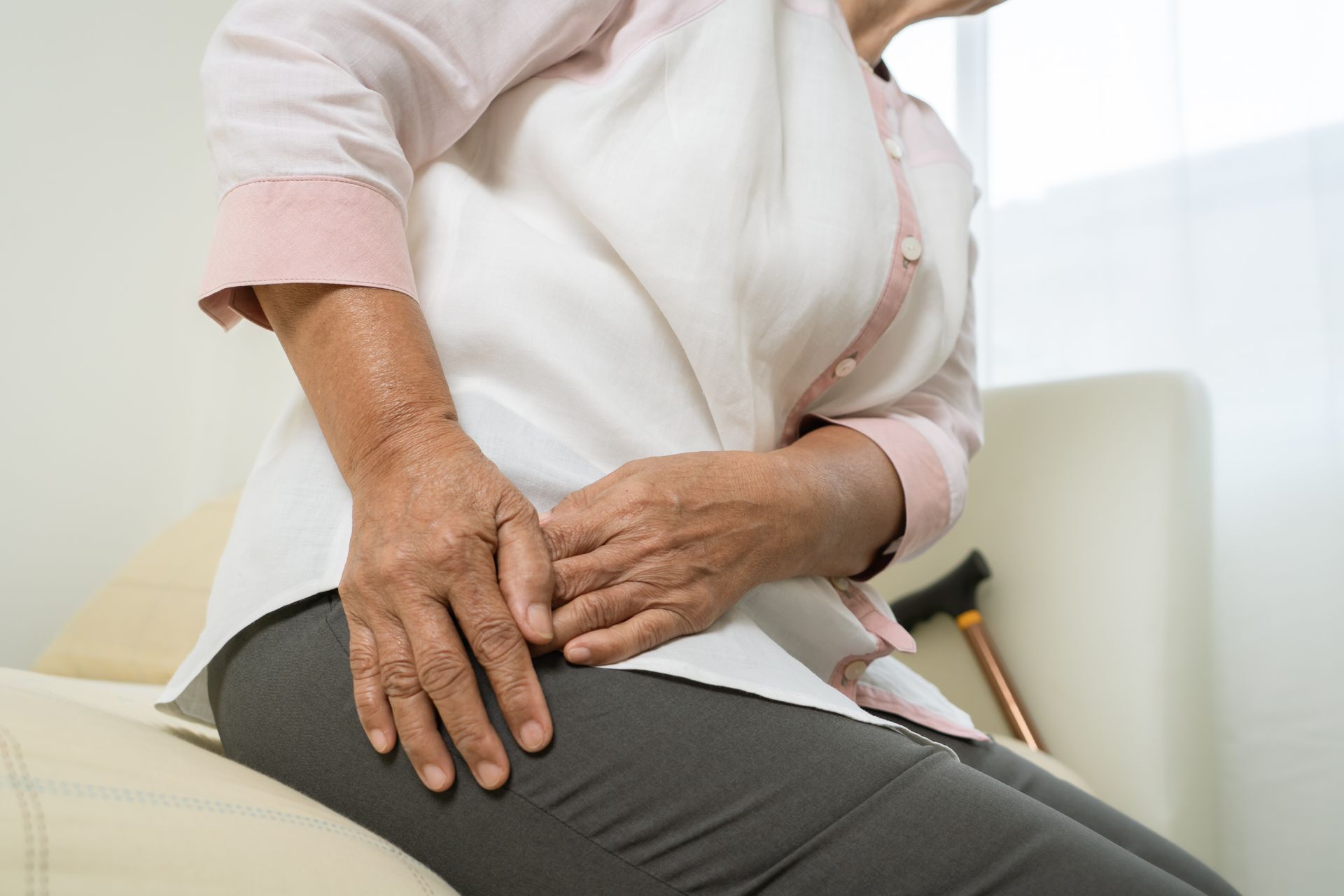 A person sits down, holding their hip with both hands, with a cane resting against a chair nearby.