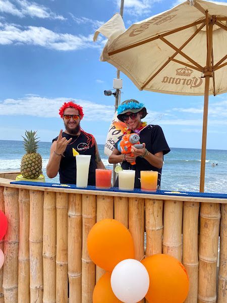 Two men are standing behind a bamboo bar with drinks and balloons.