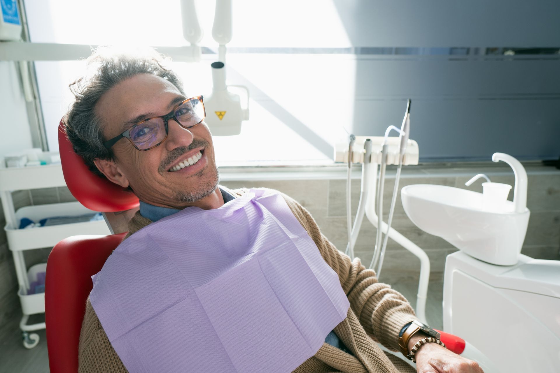Man in dental chair smiling at the camera, wearing glasses and a bib. Dental equipment in the background.