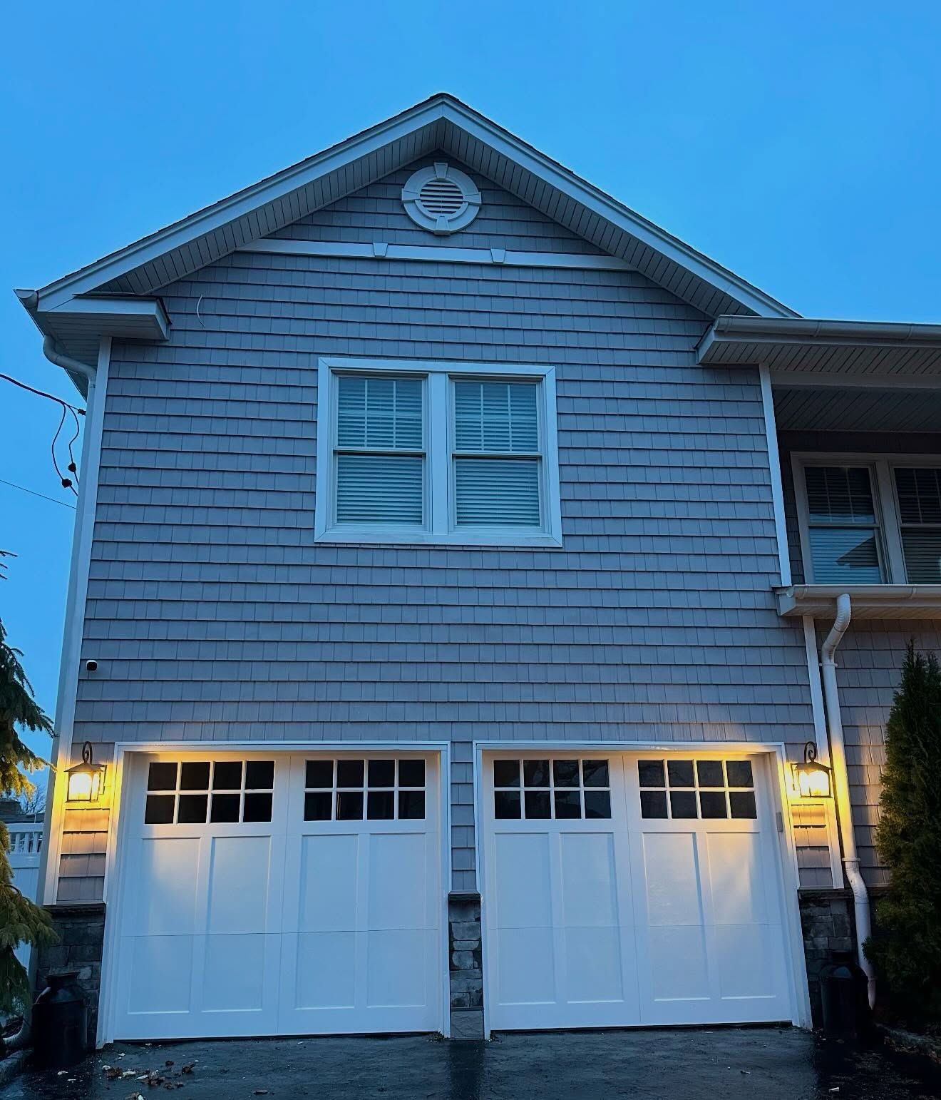Two-story gray house with white garage doors at dusk, lit by warm exterior lights