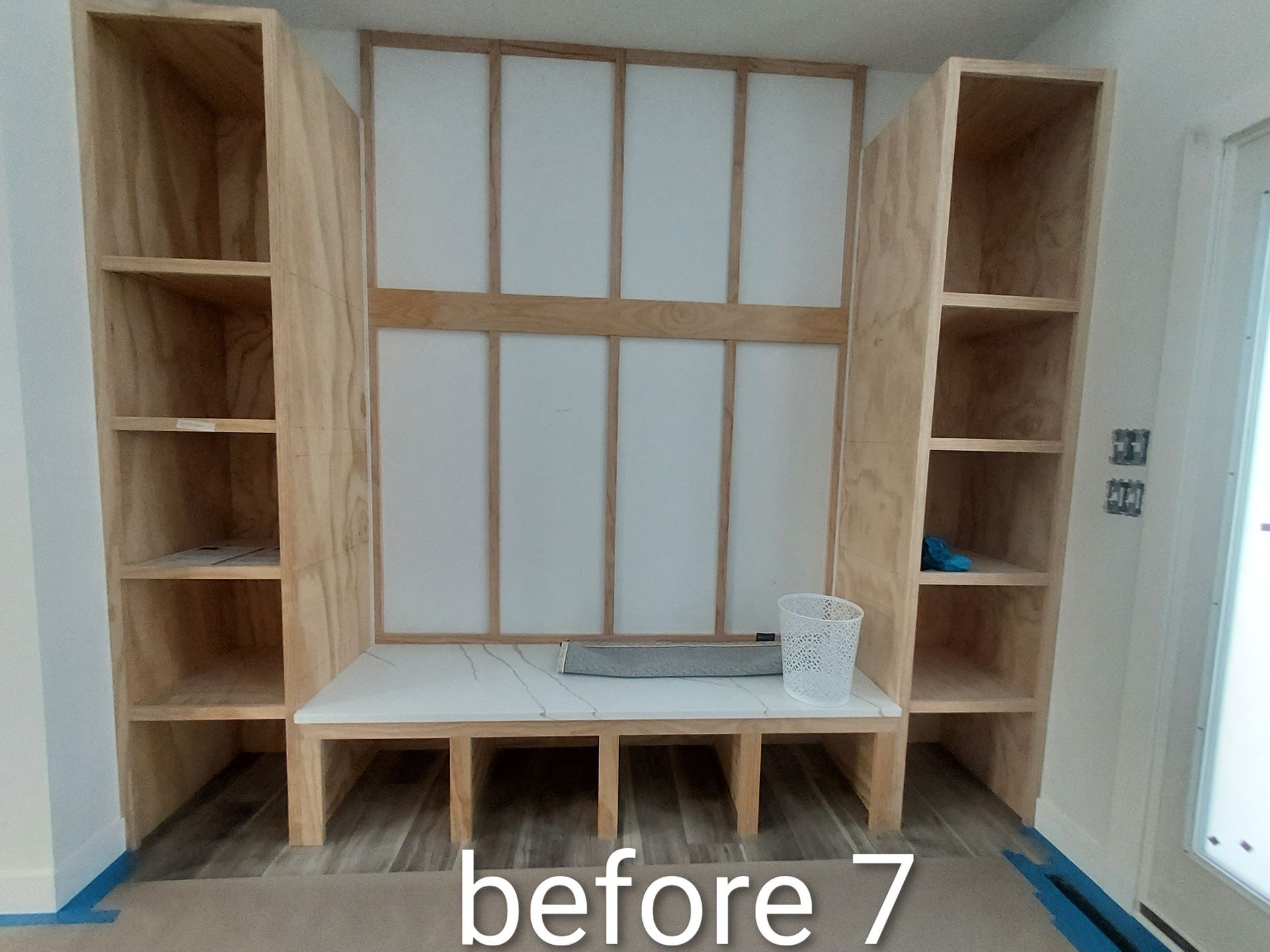 Built-in wooden shelving and desk unit in a white room, unfinished cabinet labeled “before 7”
